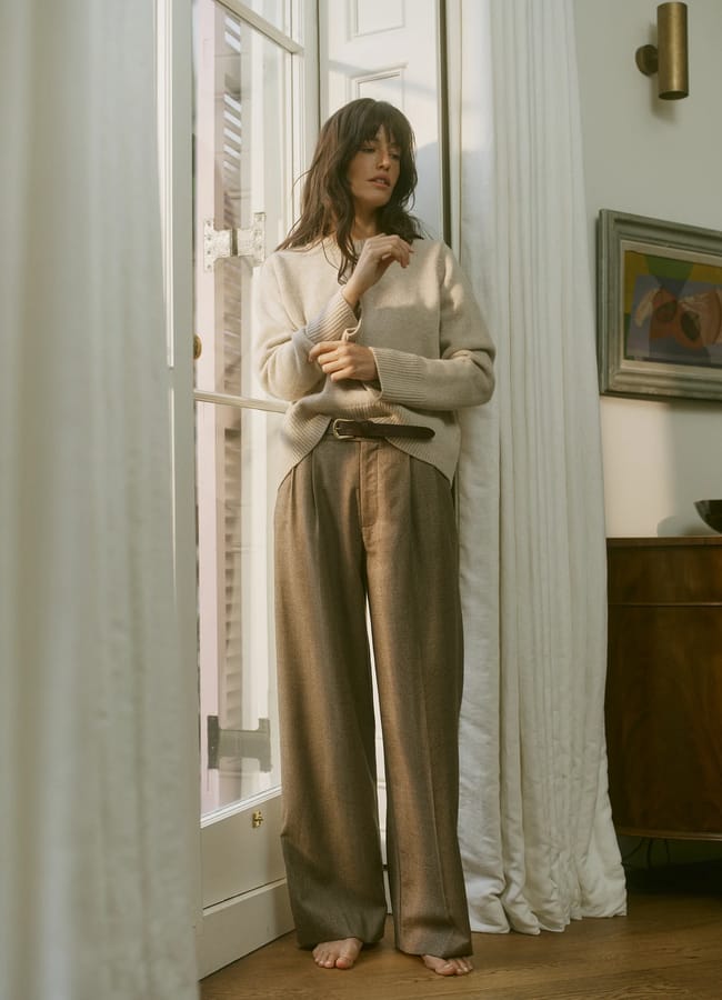 A woman with long hair stands barefoot indoors by a glass door, wearing a loose beige jumper and wide pleated brown trousers with a belt. Sunlight filters through the curtains, and framed art is visible on the wall behind her.