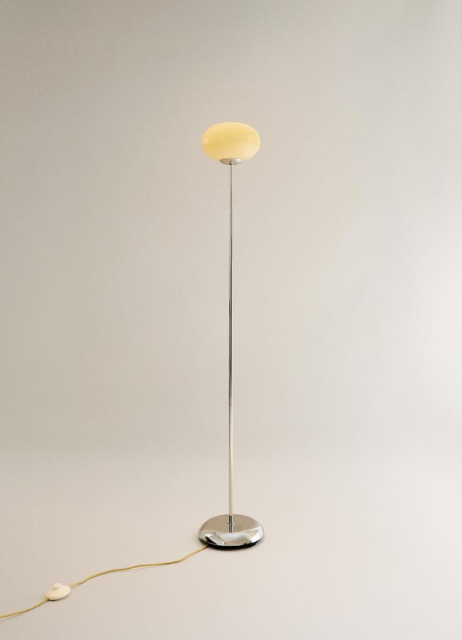 A minimalist floor lamp with a round, yellowish lampshade, slim chrome stand, and circular chrome base evokes high street homeware style. It features a power lead with a round foot switch, all set against a light background.