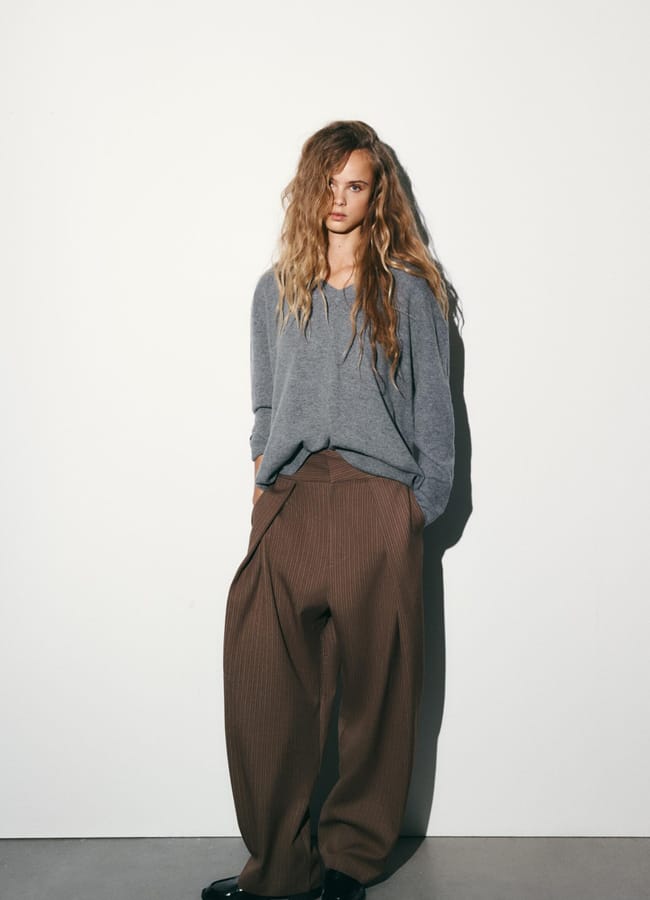 A person with long wavy hair stands against a white wall wearing a loose grey jumper, wide pleated trousers in brown, and black shoes, hands in their pockets and a relaxed posture.
