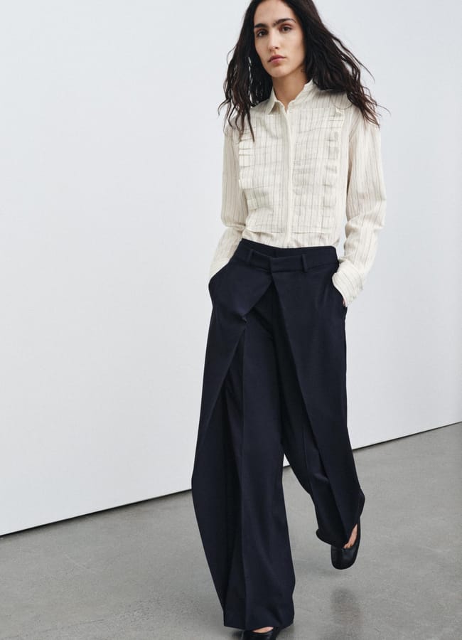 A person with long, wavy dark hair wears a cream-coloured textured blouse and loose-fitting, pleated black trousers whilst walking indoors on a concrete floor against a plain white wall.