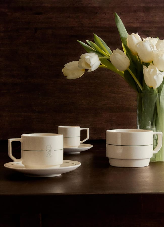 A glass vase with white tulips sits on a dark wooden table beside three high street homeware cups with matching saucers and handles, each cup featuring a simple green line design.