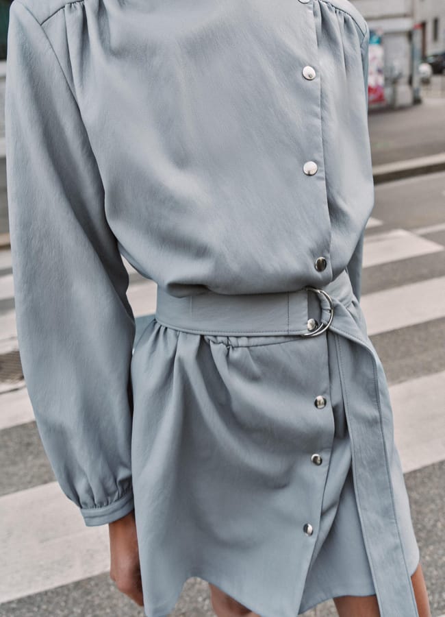 A person wearing a light grey, long-sleeved dress with silver buttons and a matching belt, standing on a zebra crossing in an urban setting—an elegant look reflecting spring summer 2026 fashion trends with its shiny texture and gathered shoulders.
