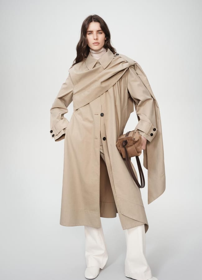 A woman with long brown hair wears a beige trench coat with an asymmetric drape, white trousers, and white shoes, holding a brown handbag against a plain light background.