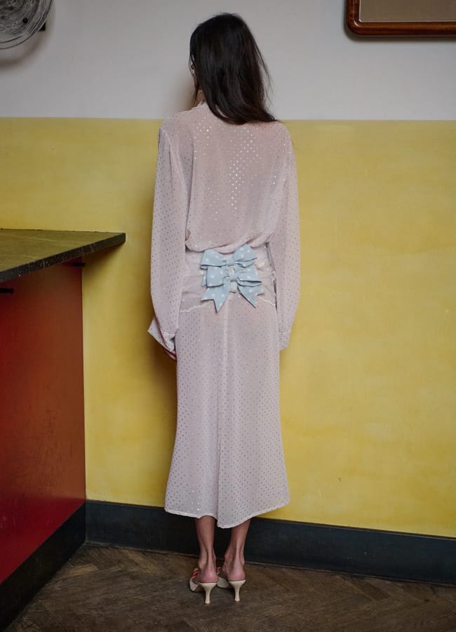 A woman stands with her back to the camera, wearing a light pink, long-sleeved, mid-length dress—highlighting spring summer 2026 fashion trends—with subtle polka dots and three light blue bows. She is in a room with yellow and red walls and dark wooden floors.