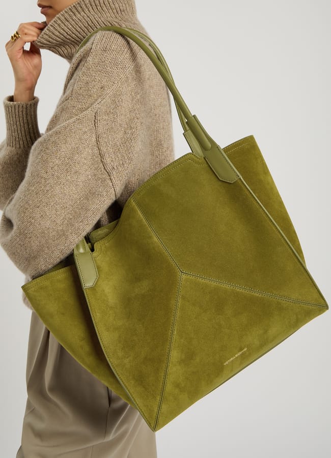 A person wearing a beige polo-neck jumper and taupe trousers carries a large olive green suede tote bag with geometric stitching and two handles over their shoulder, showing off the season’s trend for big bags.