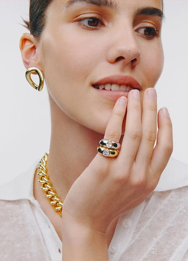 A woman wearing a sheer white collared shirt showcases gold jewellery—chunky chain necklace, looped earring, and two gold rings—as she touches her face and smiles, embodying fresh spring summer 2026 fashion trends.