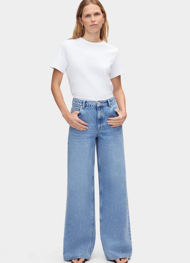 A person stands facing forwards, wearing a plain white T-shirt and high-waisted, wide-leg light blue jeans, with hands in their front pockets against a plain white background.