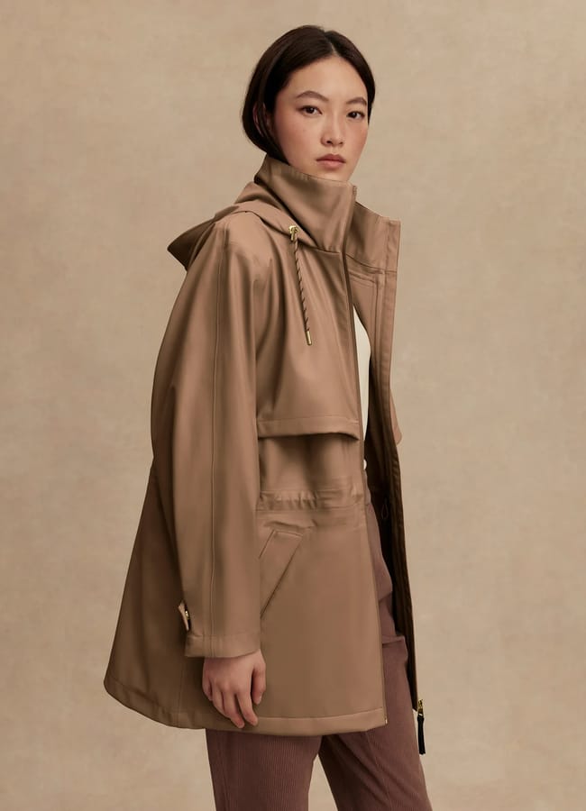 A person with straight, dark hair looks at the camera whilst wearing a stylish waterproof jacket with a hood and brown trousers, standing against a beige background.