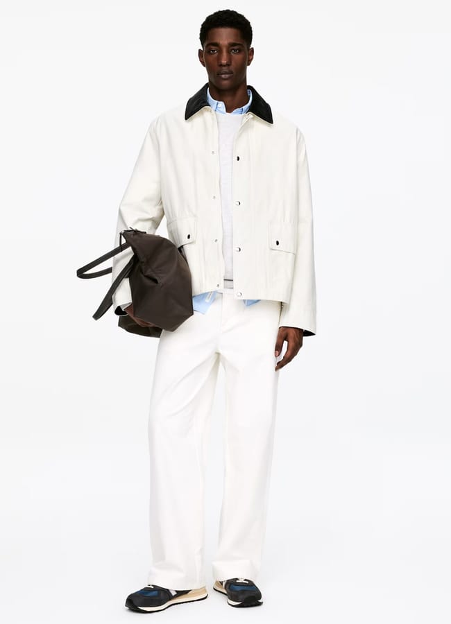 A man stands against a white background wearing a stylish light-coloured men's jacket, white wide-leg trousers, dark shoes, and holding a dark tote bag in his left hand.