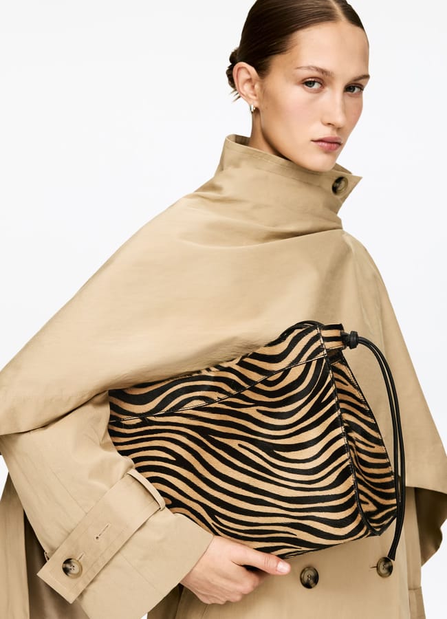 A woman in a beige trench coat holds a striking zebra print clutch bag with a black drawstring. She has her hair tied back and stands against a plain white background.