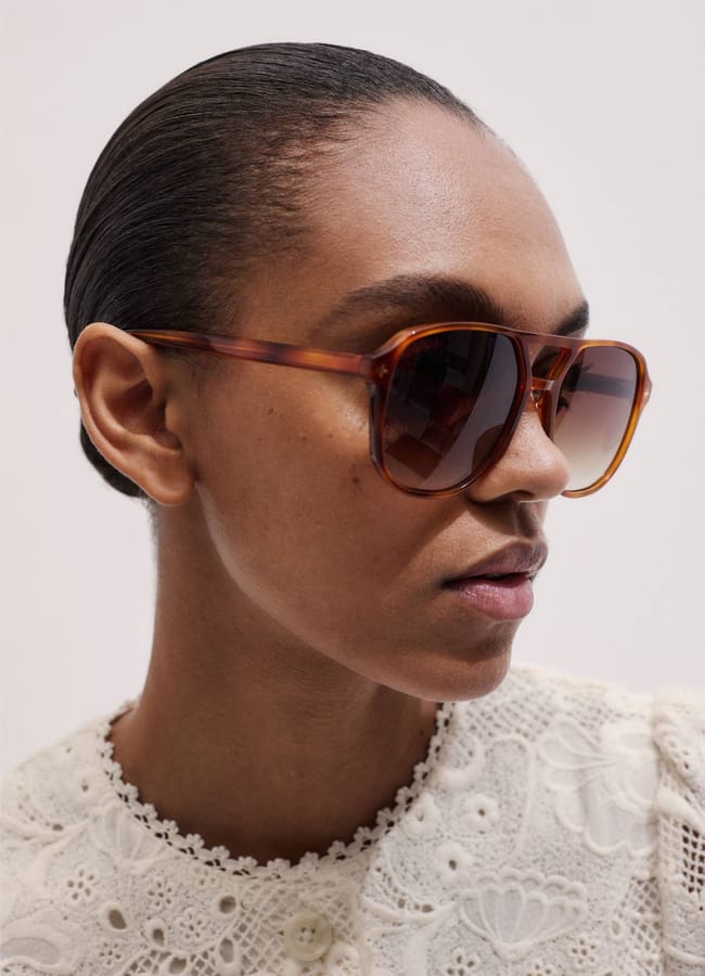 A person with slicked-back hair wears large brown sunglasses and a white lace top, looking slightly to the side against a plain background.