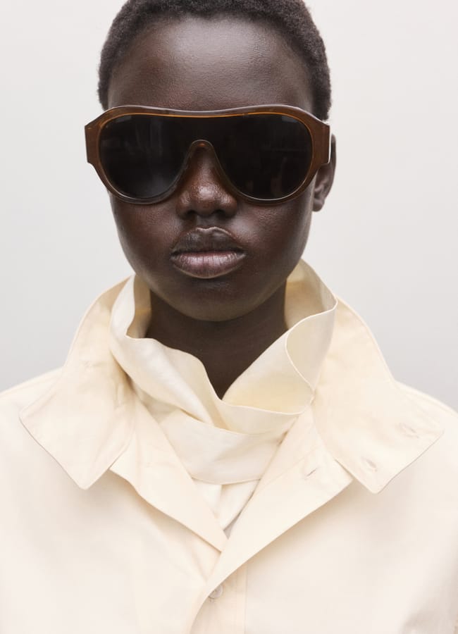 A person with dark skin wears oversized dark sunglasses, a light beige collared shirt, and a loosely wrapped matching scarf, posing against a plain, light background.
