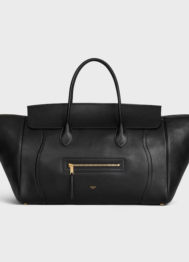 A black leather designer handbag with wide, winged sides embodies the bold style of large bags, featuring two top handles, a front zip pocket with a gold zip, and subtle curved stitching details, displayed on a plain light background.