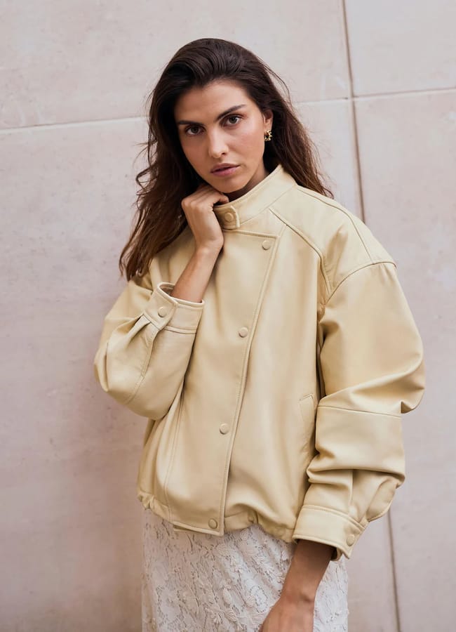A woman with long brown hair stands against a light stone wall, wearing a beige oversized jacket with press studs and a lace skirt—an effortlessly chic look inspired by spring summer 2026 fashion trends—as she gazes thoughtfully at the camera.