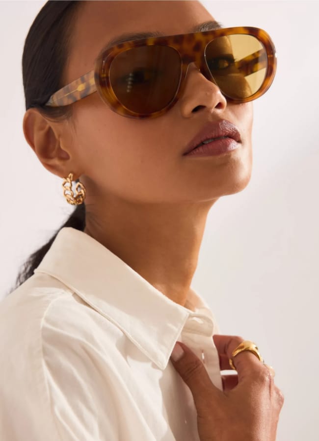 A woman wearing oversized tortoiseshell sunglasses, gold earrings, and a white collared shirt looks confidently at the camera, her hair pulled back as she adjusts her shirt collar.