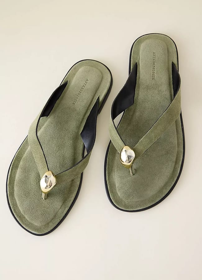 A pair of sage green suede thong sandals with black accents and a round gold decorative detail on the straps, displayed on a light-coloured surface.