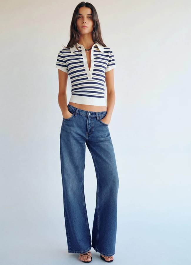 A woman stands against a plain background wearing a short-sleeved, striped polo crop top, high-waisted wide-leg blue jeans, and black sandals, with hands in her pockets and a relaxed expression.