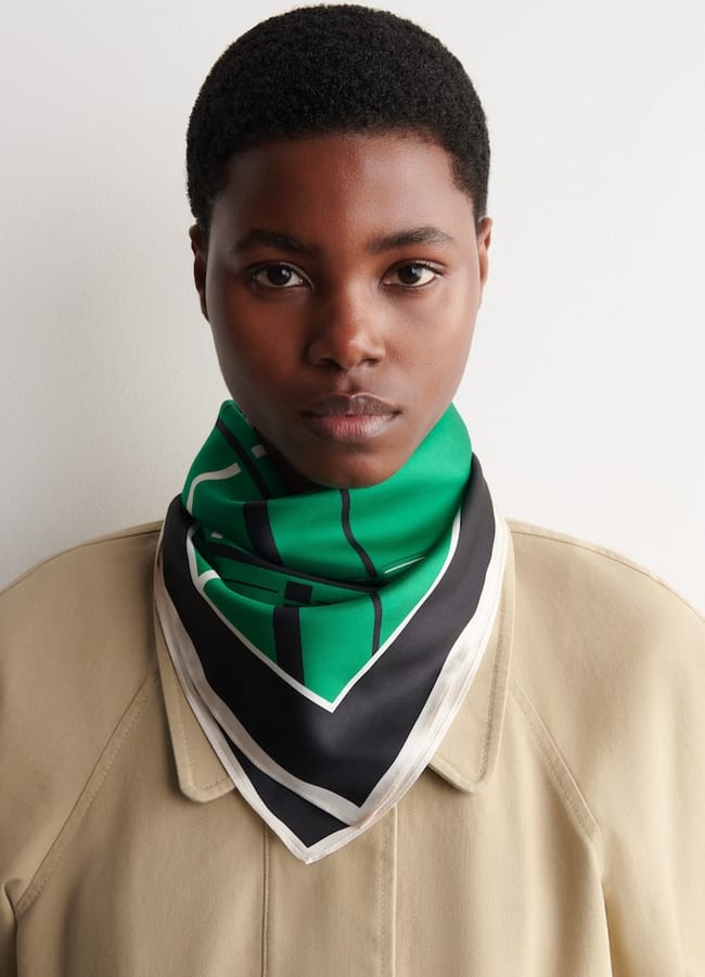 A person with short hair wears a beige coat and a green, black, and white patterned scarf around their neck, standing against a plain white background.