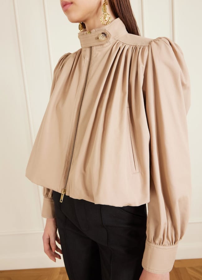 A woman wears a beige, high-neck, puff-sleeve jacket with a front zip and gold statement earrings—a chic nod to spring/summer 2026 fashion trends. The jacket features gathered shoulders and falls loosely to the waist, paired stylishly with black trousers.