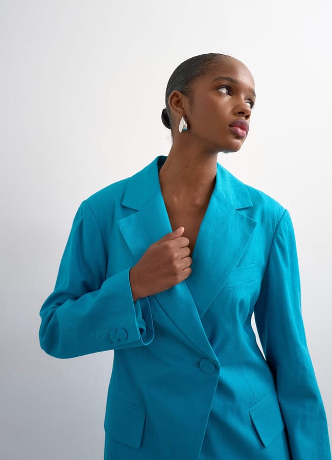 A person wearing a bright turquoise blazer, a must-have fashion piece, stands against a plain white background, looking thoughtfully to the side. They have short hair pulled back and are wearing silver drop earrings.