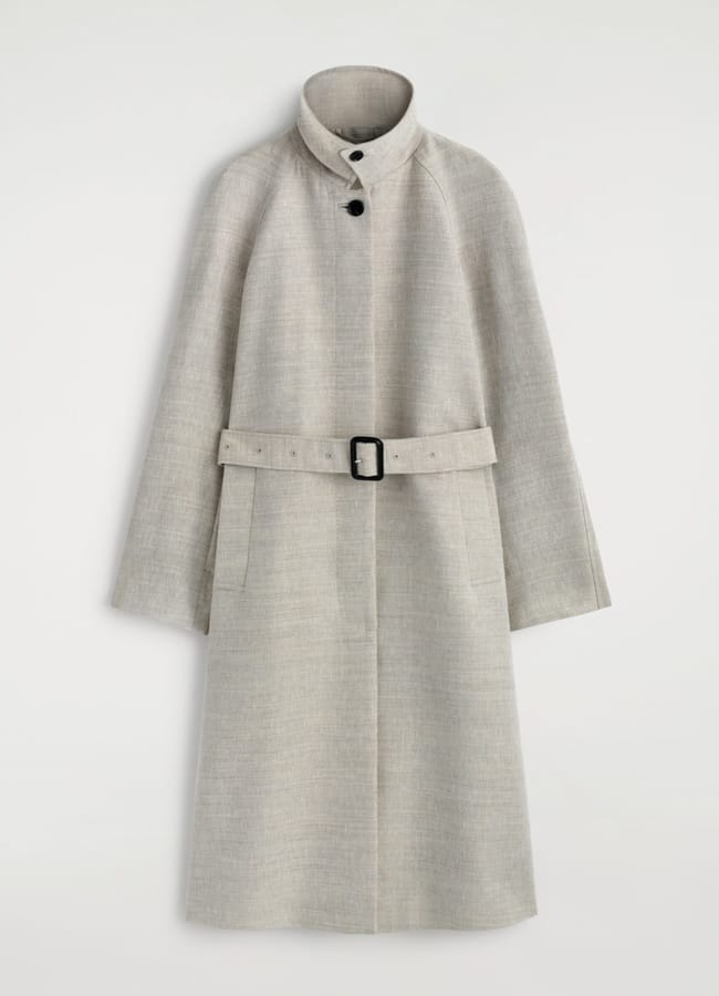 A light grey, knee-length wool coat with a high collar, black buttons near the neckline, long sleeves, front pockets, and a matching belt with a black rectangular buckle—perfect for channelling Carolyn Bessette Kennedy’s cool '90s wardrobe.