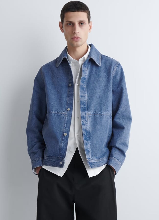 A man wearing a stylish light blue denim men's jacket over a white button-up shirt and black trousers stands against a plain white background, looking directly at the camera with his hands in his pockets.