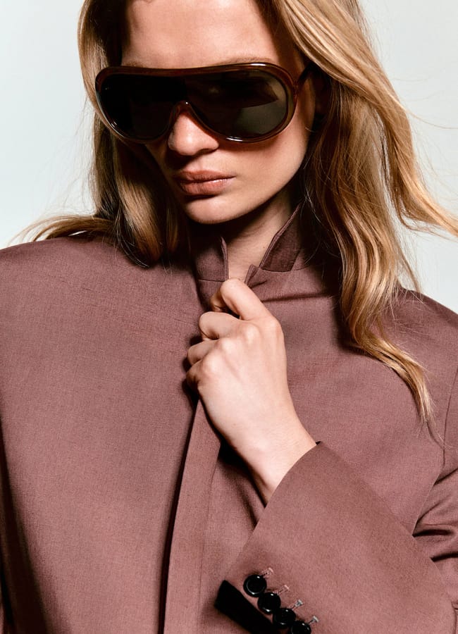 A woman with long blonde hair wears large, dark sunglasses and a mauve blazer, holding the collar with one hand. She looks serious and confident against a neutral background.