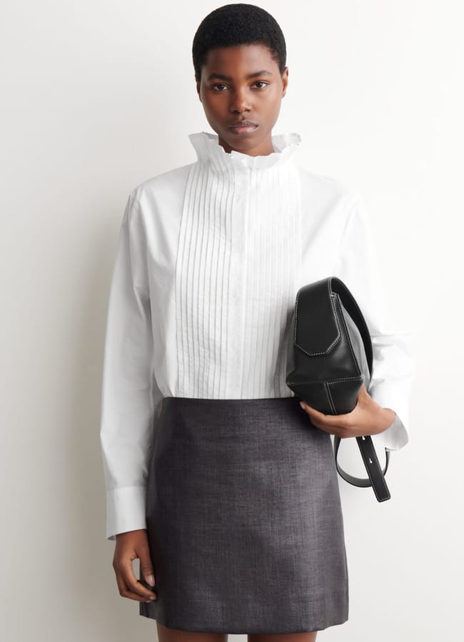 A person wearing a white pleated blouse and a dark grey skirt stands against a plain light background, holding a black handbag at their side—an effortlessly chic nod to spring/summer 2026 fashion trends.