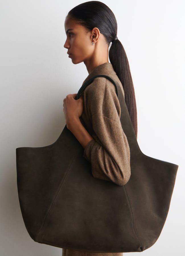 A woman with long dark hair in a low ponytail wears a brown outfit and carries one of this season’s must-have big bags—a large, dark brown suede tote—over her shoulder, standing in profile against a plain white background.