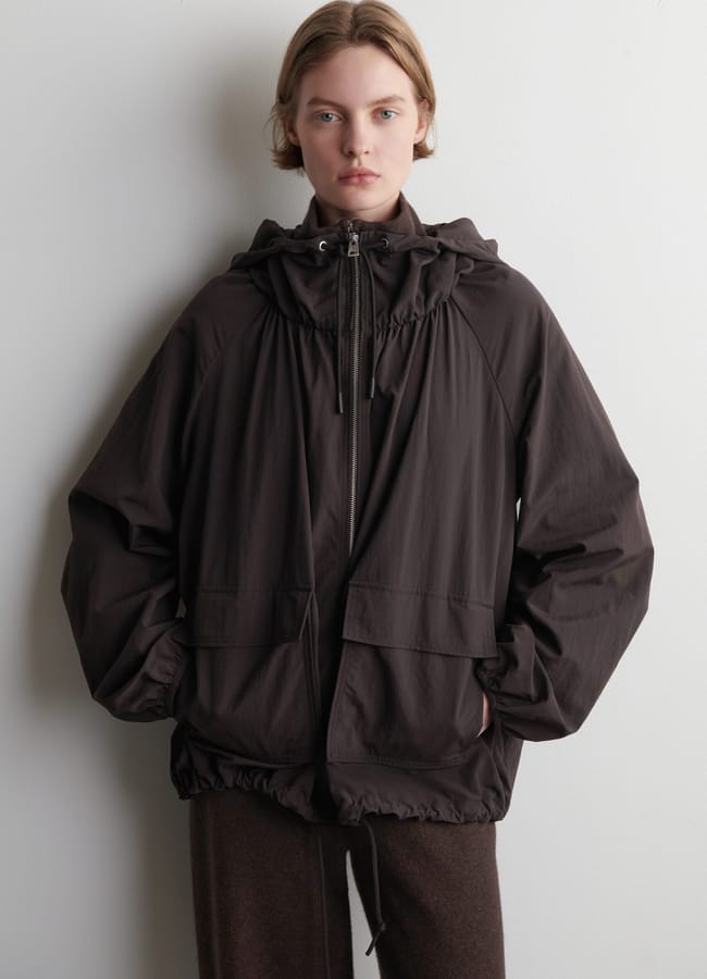 A person with short, fair hair stands against a plain white wall, wearing a loose-fitting dark brown stylish waterproof jacket with large front pockets and matching brown trousers. Their hands are in their pockets, and they have a neutral expression.