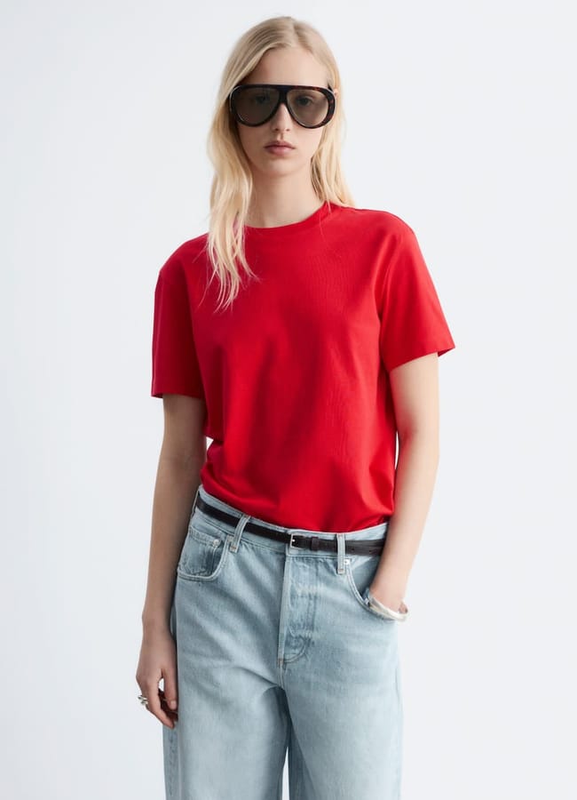 A person with long blonde hair wearing oversized black sunglasses, a bright red T-shirt, and light blue jeans stands against a plain white background with one hand in their pocket.