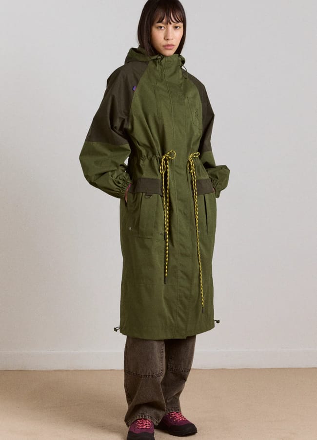 A person stands indoors wearing a stylish waterproof jacket—a long green hooded parka with yellow drawstrings—paired with brown trousers and pink and black shoes. The background features a light-coloured wall and plain carpet.