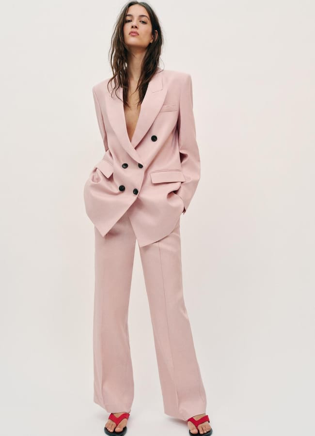 A woman stands against a plain background wearing an oversized light pink suit—this season's fashion must-have—paired with red heeled sandals. Her hands are in her pockets, her long hair is loose, embodying effortless confidence worthy of a Style Editor.