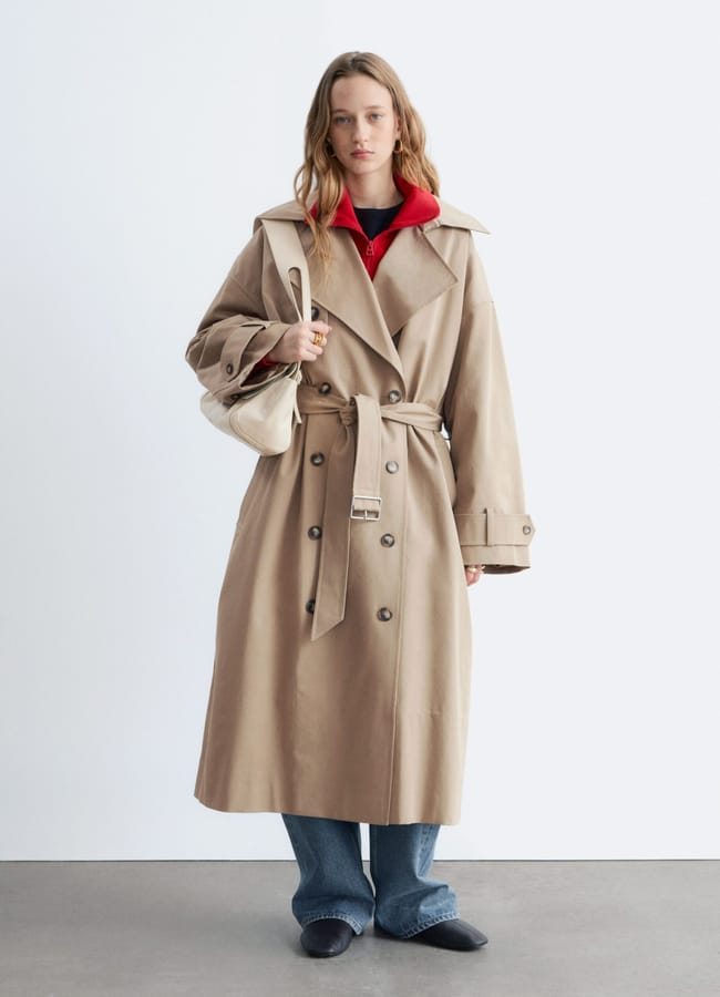 A person wearing a long beige trench coat, blue jeans, and black shoes stands against a plain white background. They have a red jumper underneath the coat and are carrying a small white shoulder bag.