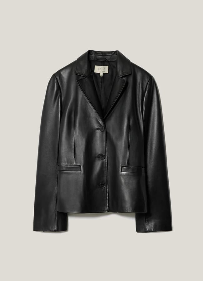 A black faux leather blazer with a notched collar, two front buttons, and two flap pockets features subtle sheer panels, displayed against a plain light grey background.
