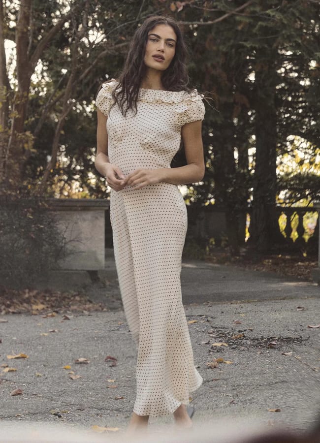 A woman with long dark hair stands outdoors on a paved path, wearing a light-coloured, ankle-length dress with a subtle pattern—an elegant nod to spring summer 2026 fashion trends. Trees and stone railings are visible in the background.