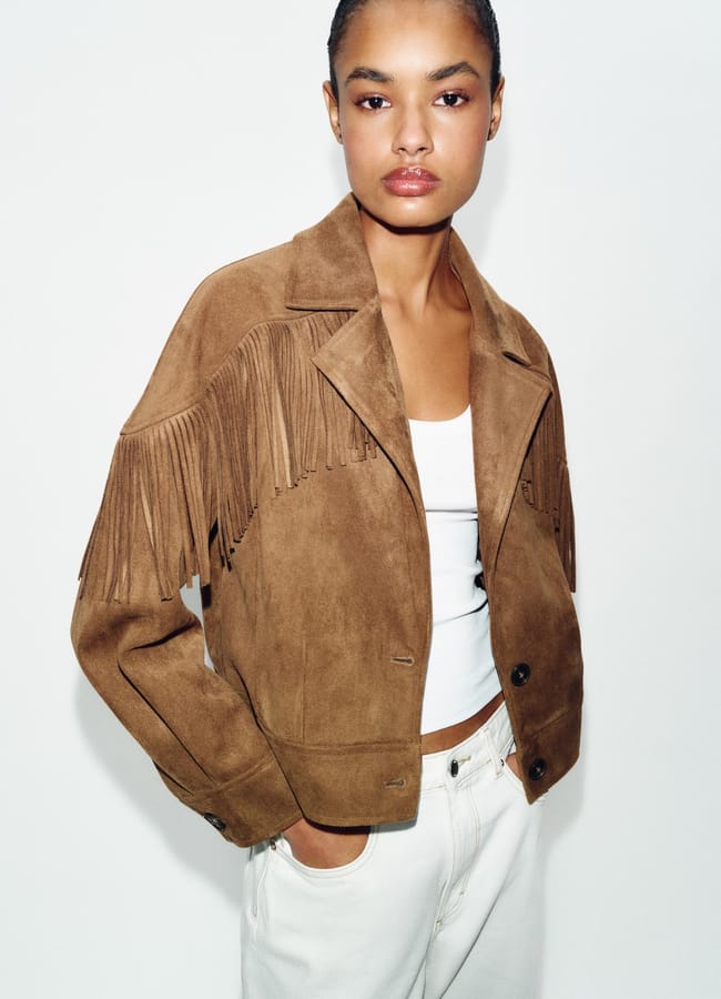 A person wearing a brown suede jacket with fringe details over a white vest and white trousers stands against a plain white background, effortlessly showcasing style that highlights the appeal of fashion’s unique fringe benefits.