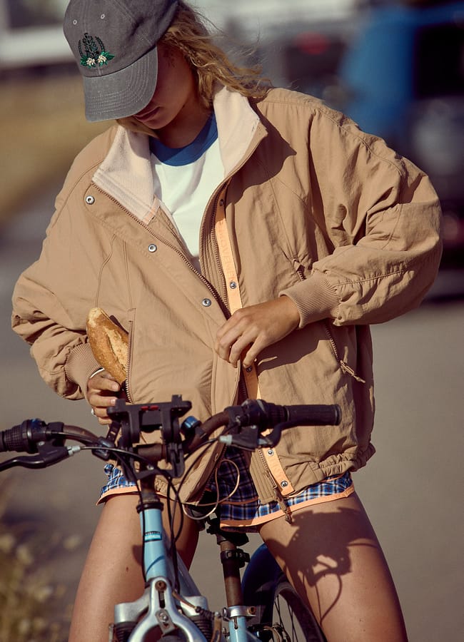 A person in a stylish waterproof jacket, checked shorts, and a grey cap stands outdoors with a blue bicycle, holding a piece of bread and adjusting their jacket. Their head is slightly bowed, hiding their face under the cap.