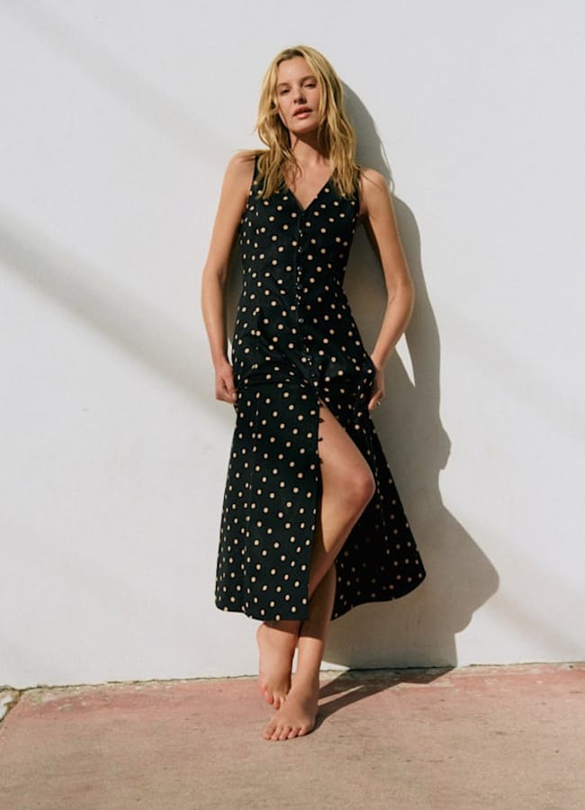 A woman stands barefoot on a sunlit pavement against a white wall, wearing a sleeveless black dress with tan polka dots and a front slit, with her hair loose and hands resting at her sides.
