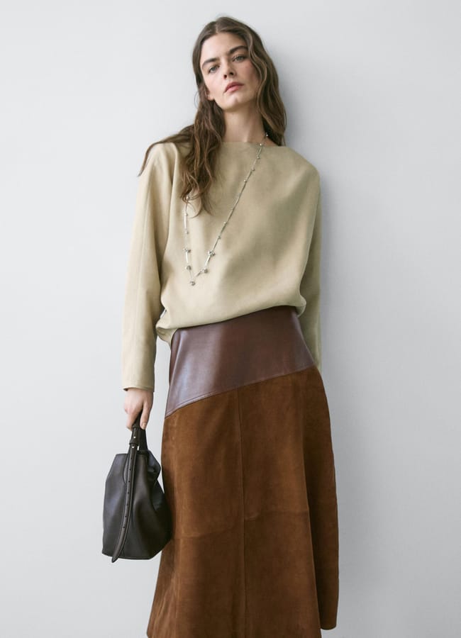A woman with long brown hair wears a beige long-sleeve top, a brown leather and suede skirt, and holds a dark handbag—capturing the calm elegance of spring/summer 2026 fashion trends against a plain light grey wall.