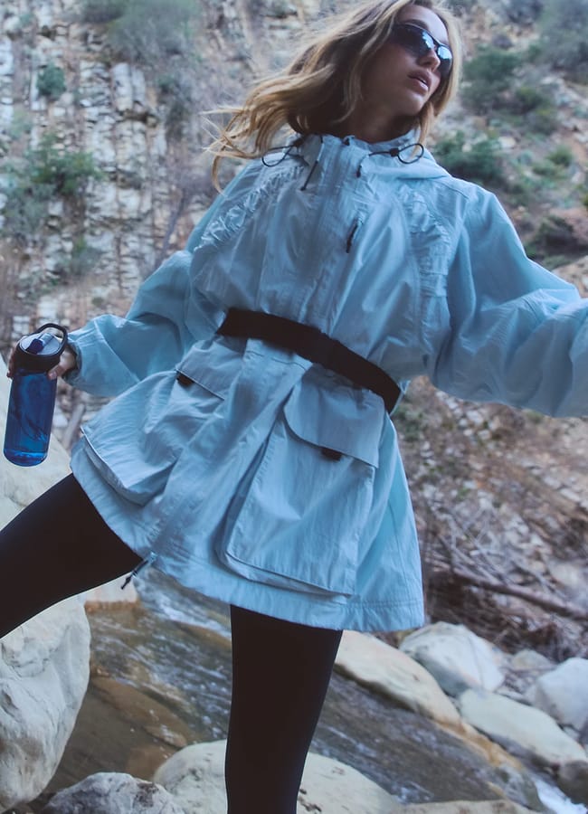 A person wearing a stylish waterproof jacket, black leggings, and sunglasses stands outdoors near rocks and a stream, holding a blue water bottle. Rocky cliffs and trees are visible in the background.