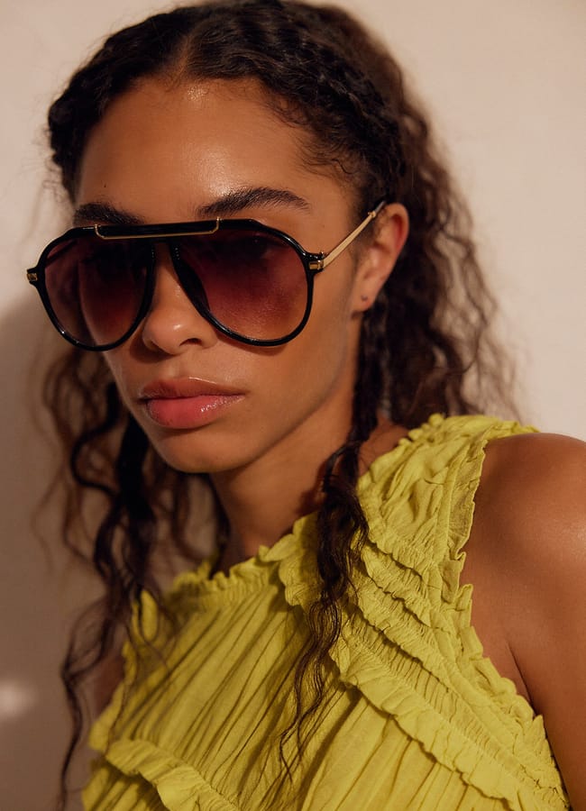 A woman with curly hair wearing large, dark aviator sunglasses and a sleeveless, frilled yellow top, looking slightly to the side against a neutral background.