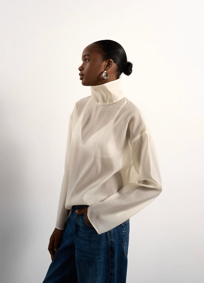 A woman in a white polo-neck blouse and blue jeans stands against a plain light background, looking to her left. Her hair is tied back, and she wears large hoop earrings—a chic wardrobe look you can easily recreate with insta dupes.