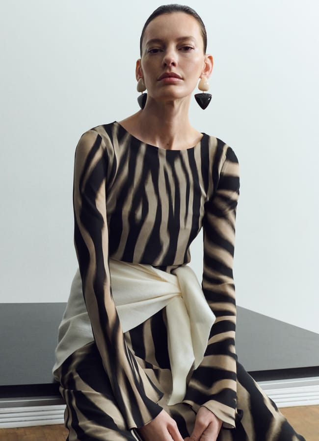 A woman with slicked-back hair wears a striking zebra-print dress, large triangular earrings, and a white jumper tied around her waist, seated against a minimalist background.