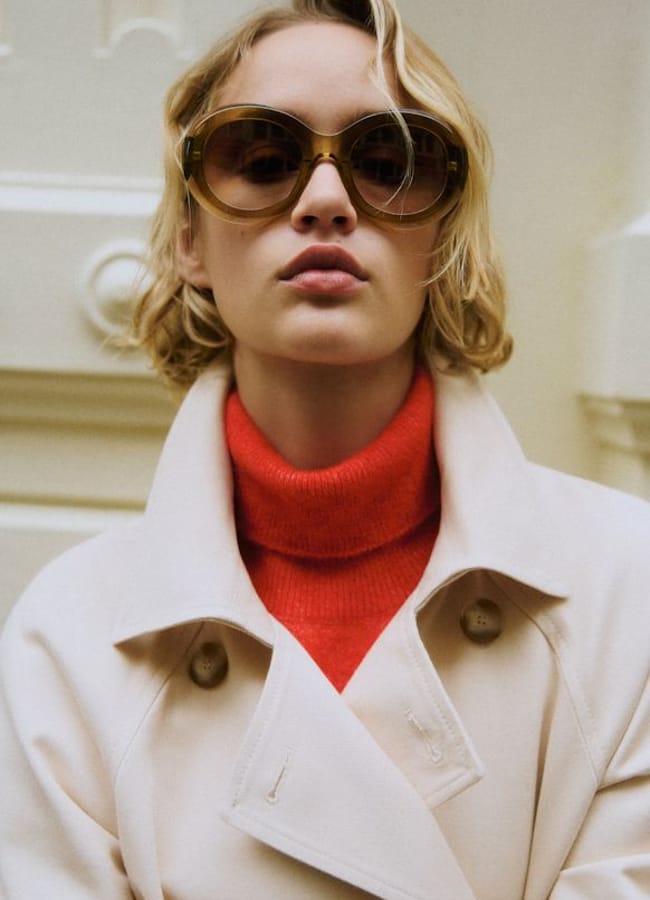A person with short blonde hair wears large round sunglasses, a beige trench coat, and a bright red polo neck, posing in front of a cream-coloured background.