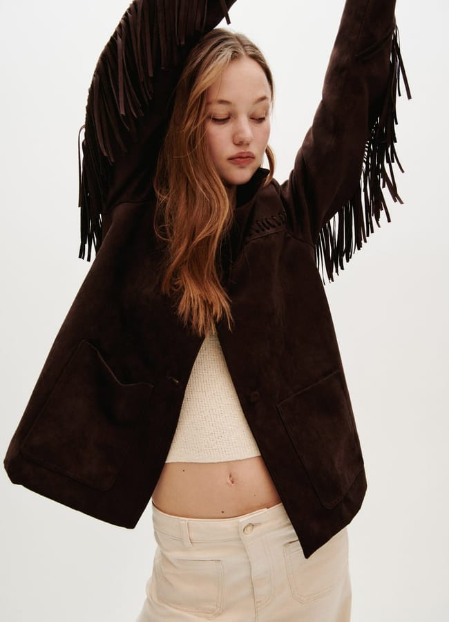 A young woman with long hair raises her arms, wearing a dark brown jacket with fringe on the sleeves, a cream knitted top, and light-coloured jeans against a plain background.