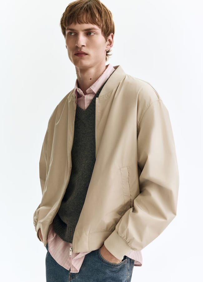A young person with short light brown hair wears a beige men's jacket over a grey jumper and pink collared shirt, paired with blue jeans, standing against a plain white background.