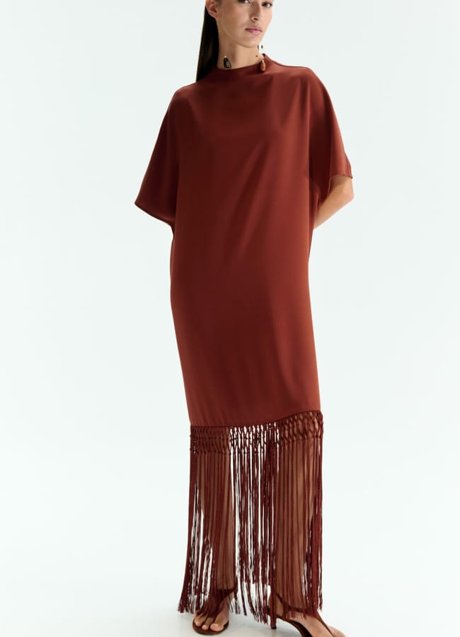 A woman wearing a loose, rust-coloured dress with short sleeves and long, fringe detailing at the hem stands against a plain white background—showcasing true fringe benefits in style. She has straight dark hair, minimal jewellery, and wears flat sandals.