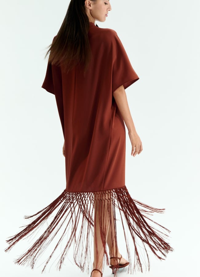 A woman wearing a rust-coloured dress with short sleeves and long fringe benefits-inspired details at the hem stands against a white background, facing away and slightly to the side. She is also wearing brown sandals.