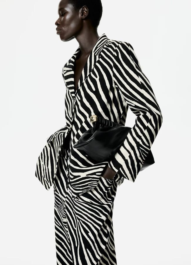 A person wearing a striking zebra print suit stands in profile, holding a black clutch bag against a plain white background.
